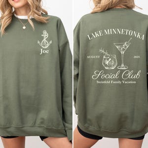 May include: Olive green sweatshirt with white graphic designs. The front features an anchor and the name "Joe." The back displays "Lake Minnetonka Social Club" with cocktail illustrations, the month of August, and the year 2025. Steinfield Family Vacation is also printed on the back.