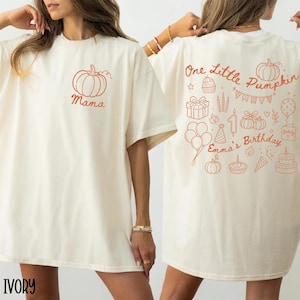 Pode incluir: Uma t-shirt cor de marfim com um pequeno gráfico de abóbora laranja e a palavra Mama no peito esquerdo. As costas exibem desenhos de linhas laranja, incluindo abóboras, um presente, balões, uma vela com o número um e o texto One Little Pumpkin Emma's Birthday.