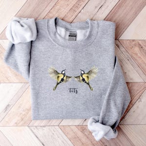 May include: Gray sweatshirt with a graphic of two yellow birds with black wings and the word "tits" printed below them.