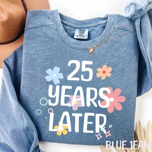 May include: A blue jean sweatshirt with the text "25 years later" in white lettering and colourful flowers surrounding the text.