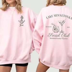 May include: Pink sweatshirt with an anchor graphic and the name "Joe" on the front. The back features text that reads "Lake Minnetonka Social Club August 2025" with cocktail illustrations. A casual, comfortable garment.