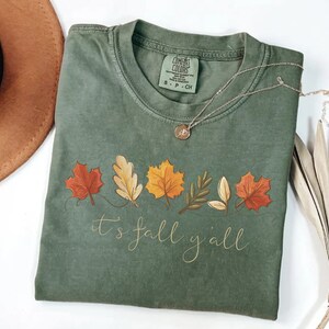 May include: Sage green Comfort Colors t-shirt with a fall leaves design and the text "it's fall y'all" in a cursive font. A silver necklace with a pendant and a brown hat are also visible.