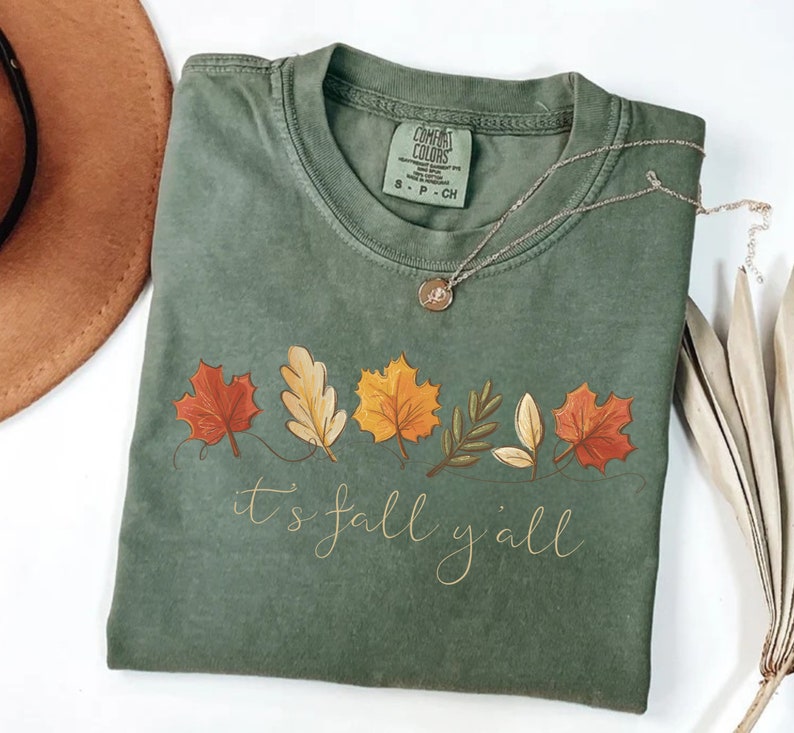 May include: Sage green Comfort Colors t-shirt with a fall leaves design and the text "it's fall y'all" in a cursive font. A silver necklace with a pendant and a brown hat are also visible.