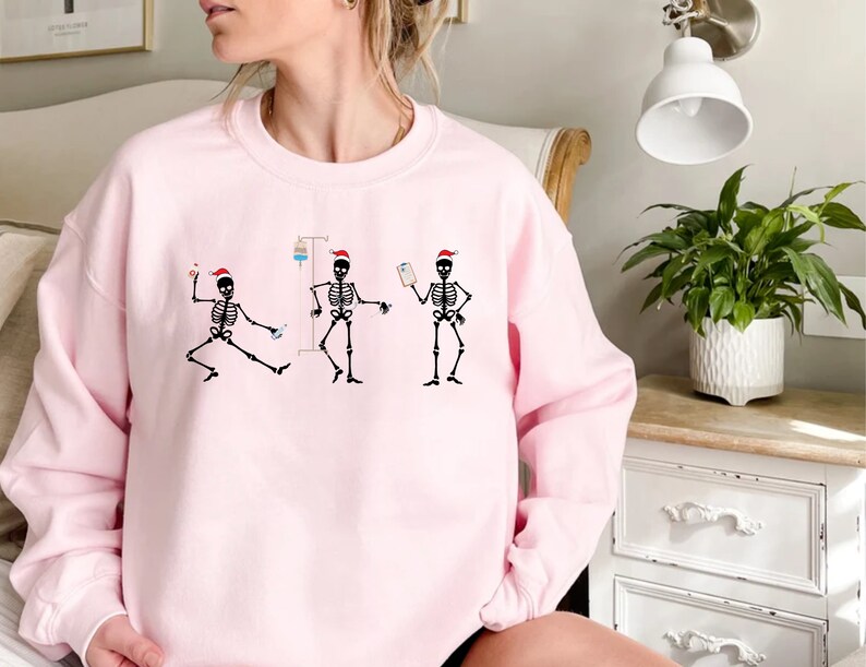 Pode incluir: Um moletom rosa claro com um desenho de tr&ecirc;s figuras de esqueleto pretas usando chap&eacute;us de Papai Noel. Cada esqueleto est&aacute; em uma pose diferente, com equipamentos m&eacute;dicos. O design &eacute; uma vers&atilde;o humor&iacute;stica dos cuidados de sa&uacute;de durante a &eacute;poca natal&iacute;cia.