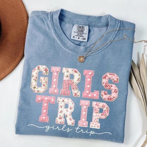 Może przedstawiać: Niebieska koszulka z krótkim rękawem, okrągłym dekoltem i metką Comfort Colors. Z przodu znajduje się napis GIRLS TRIP wykonany z dużych, naszywanych liter z tkaniny w kwiatowe, kratkowane i kropkowane wzory, a poniżej napis girls trip pisany kursywą.