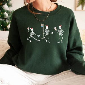 Pode incluir: Camisola verde escuro com um design de tr&ecirc;s figuras de esqueleto branco usando chap&eacute;us de Papai Noel. Os esqueletos s&atilde;o retratados em v&aacute;rias poses, um segurando uma seringa, outro com um gotejamento intravenoso e o terceiro segurando uma carta. Um design festivo e divertido.