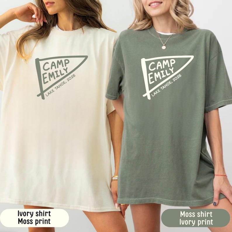 May include: Two short-sleeve crew neck t-shirts. The left shirt is ivory with a moss green graphic of a pennant flag featuring the text CAMP EMILY and LAKE TAHOE, 2026. The right shirt is moss green with an ivory graphic of the same design.