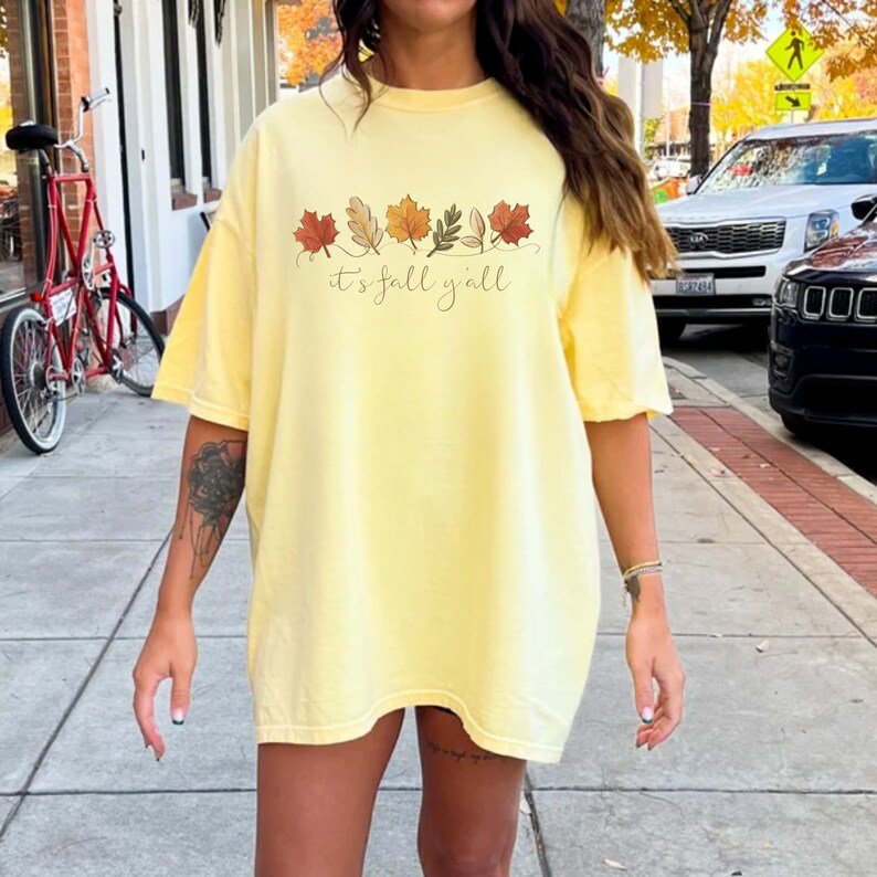 May include: A light yellow t-shirt with the phrase "it's fall y'all" in a script font. Above the text is a row of stylized fall leaves in shades of red, orange, and brown. The shirt has short sleeves and a relaxed fit.