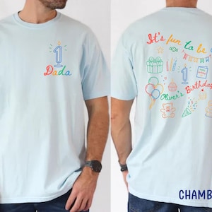 May include: Light blue t-shirt with colorful text and graphics. The front features a candle with the word "Dada". The back displays the phrase "It's fun to be one" with birthday-themed illustrations, including balloons, a cake, and the name "Oliver's Birthday".