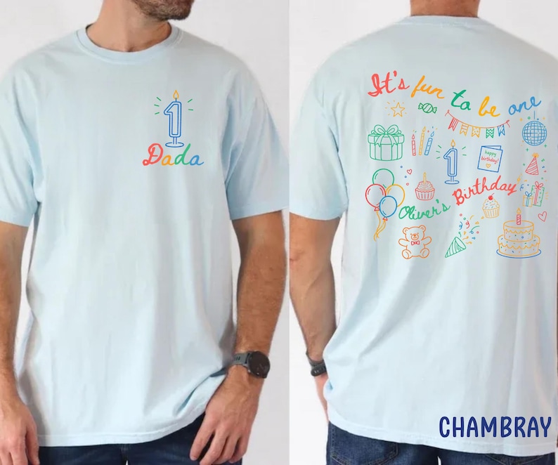 May include: Light blue t-shirt with colorful text and graphics. The front features a candle with the word "Dada". The back displays the phrase "It's fun to be one" with birthday-themed illustrations, including balloons, a cake, and the name "Oliver's Birthday".