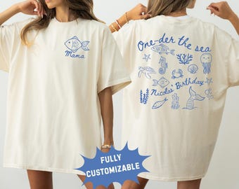 May include: An off-white t-shirt featuring blue line art. The front displays a fish and the word "Mama." The back shows various sea creatures, including a jellyfish, crab, and seahorse, with the text "One-der the sea" and "Nicolas' Birthday."