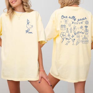 Peut inclure: T-shirt jaune clair avec un motif d'oie bleue et le mot "Mom" sur le devant. Le dos du t-shirt pr&eacute;sente le texte "One silly goose" et des illustrations sur le th&egrave;me de l'anniversaire, dont un g&acirc;teau et des ballons. Style d&eacute;contract&eacute;.