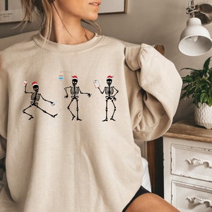 Pode incluir: Camisola bege com um gr&aacute;fico de tr&ecirc;s esqueletos dan&ccedil;ando usando chap&eacute;us de Papai Noel. Cada esqueleto &eacute; retratado com equipamentos m&eacute;dicos, incluindo um gotejamento intravenoso e uma prancheta. O design &eacute; em preto e vermelho.