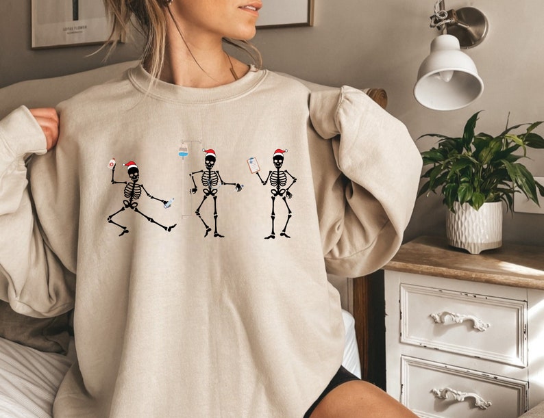 Pode incluir: Camisola bege com um gr&aacute;fico de tr&ecirc;s esqueletos dan&ccedil;ando usando chap&eacute;us de Papai Noel. Cada esqueleto &eacute; retratado com equipamentos m&eacute;dicos, incluindo um gotejamento intravenoso e uma prancheta. O design &eacute; em preto e vermelho.