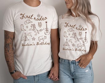 May include: Two white t-shirts with a brown graphic design of a cowboy hat, boots, a horse, a guitar, a tent, and fireworks. The text on the shirts reads "First Rodeo" and "Hudson's Birthday".