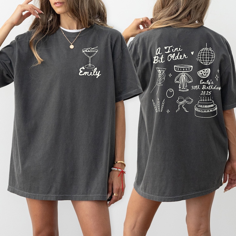 May include: Dark gray t-shirt with white graphic designs. The front features a cocktail glass and the name "Emily." The back displays the text "A Tiny Bit Older" with birthday-themed illustrations, including a disco ball and cake. The shirt is a casual, unisex style.