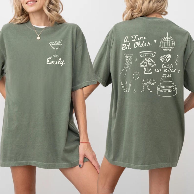 May include: Olive green t-shirt with white graphic of a martini glass and the name "Emily". The back of the shirt features the text "A Tiny Bit Older" and birthday-themed illustrations, including a disco ball and cake.