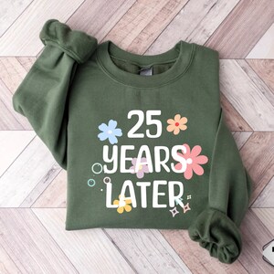 May include: A green sweatshirt with the text "25 years later" in white lettering. The text is surrounded by colourful flowers and sparkles.
