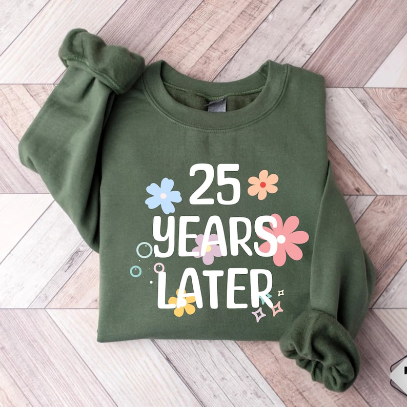May include: A green sweatshirt with the text "25 years later" in white lettering. The text is surrounded by colourful flowers and sparkles.