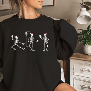 Pode incluir: Camisola preta com um gr&aacute;fico de tr&ecirc;s figuras de esqueleto usando chap&eacute;us de Papai Noel. Os esqueletos s&atilde;o retratados em v&aacute;rias poses, um dan&ccedil;ando, um com um gotejamento intravenoso e outro segurando um gr&aacute;fico. O design &eacute; branco.