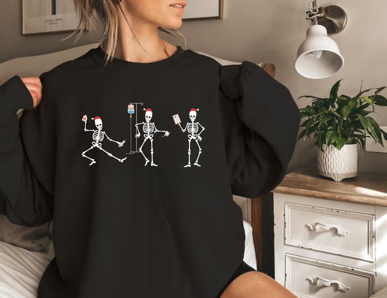 Pode incluir: Camisola preta com um gr&aacute;fico de tr&ecirc;s figuras de esqueleto usando chap&eacute;us de Papai Noel. Os esqueletos s&atilde;o retratados em v&aacute;rias poses, um dan&ccedil;ando, um com um gotejamento intravenoso e outro segurando um gr&aacute;fico. O design &eacute; branco.