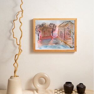 Puede incluir: Una pintura de acuarela enmarcada de dos casas pequeñas con un cielo rosa y azul. Las casas están rodeadas de árboles y un cuerpo de agua. La pintura está en una pared blanca con un jarrón de ramas y dos pequeños jarrones de cerámica sobre una mesa.
