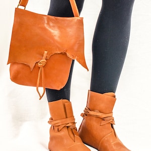 May include: A pair of brown leather ankle boots with laces tied around the ankles. The boots are worn by a person wearing black tights and a brown leather satchel bag with a strap.