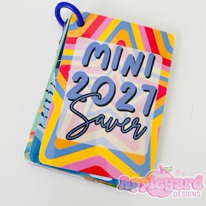 May include: A small, rectangular booklet bound with a blue plastic ring. The cover features a vibrant, multi-coloured star pattern in shades of yellow, orange, pink, and blue. The text reads MINI 2021 Saver in bold, dark blue lettering.
