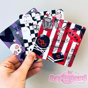 May include: Three rectangular cards featuring graphic illustrations. One card has a purple and black striped background with a skull. Another has a black and white checkerboard pattern with ghosts. The third has red and white stripes with skeletons, hearts, and the text LOVE YOU TO DEATH.