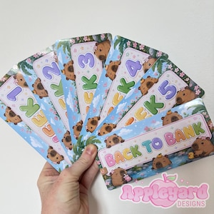 May include: A set of rectangular paper trackers featuring cartoon capybaras, floral accents, and blue sky backgrounds. The cards are labelled WEEK 1 through WEEK 5, with one card titled BACK TO BANK. Each card displays colourful text and small illustrations.