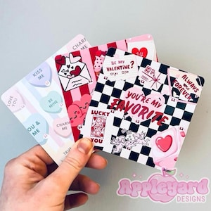 May include: Three square cards featuring Valentine's Day themes. One card has pastel heart candies with text like KISS ME and BE MINE. Another has red stripes and hearts. The third has a black and white checkerboard pattern with text YOU'RE MY FAVORITE.