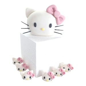 May include: A white Hello Kitty head with a pink bow, black whiskers, and black eyes sits on a white block. Several smaller Hello Kitty heads with pink bows are arranged around the base. The design is simple and cartoonish.