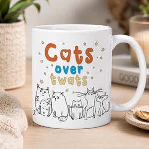 Op de afbeelding: Witte keramische mok met de tekst "Cats over twats" in oranje, blauw en geel. De mok heeft een lijntekening van zes katten en kleine sterillustraties. Een gouden lepel ligt op een klein bord.