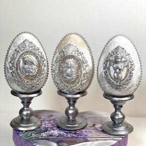 Op de afbeelding: Drie zilveren decoratieve eieren met sierlijke ontwerpen en reliëfs van religieuze figuren. Elk ei staat op een zilveren standaard.