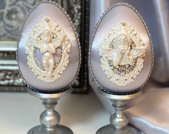 Puede incluir: Dos huevos decorativos con un soporte plateado. Cada huevo está cubierto de una tela de color morado claro con un diseño de ángel blanco con cuentas. Los ángeles tienen las manos juntas en oración.