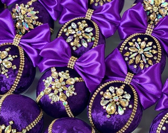 May include: Purple velvet Christmas ornaments with gold trim and jeweled embellishments. The ornaments are decorated with a large bow made of purple satin ribbon.