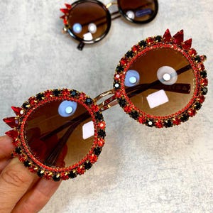 May include: Round sunglasses with a gold frame and red and black rhinestones. The sunglasses have brown lenses.