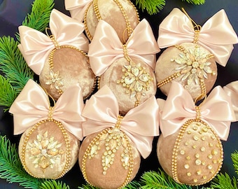 May include: A set of seven blush-colored velvet Christmas ornaments with gold trim and sparkling rhinestones. Each ornament features a pink satin bow.