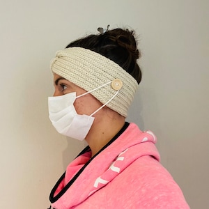 Könnte beinhalten: Ein weißes Strick Stirnband mit einem Knopf an der Seite zum Befestigen einer Gesichtsmaske. Das Stirnband wird von einer Person mit einer weißen Gesichtsmaske getragen.