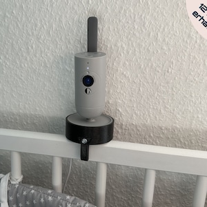 Może przedstawiać: Szary monitor dla dziecka z obiektywem kamery zamontowanym na czarnym stojaku. Stojak jest przymocowany do białej szczebelkowej barierki łóżeczka za pomocą paska. W prawym górnym rogu obrazu widoczny jest tekst "In 16 Farben erhältlich".