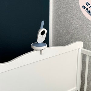 Könnte beinhalten: Ein weißer Babymonitor mit einem blauen Kamerasensor ist an einem weißen Babybett befestigt. Der Monitor ist mit einem weißen Clip am Babybett befestigt. Das Babybett ist aus weißem Holz und hat ein weißes Lattenrost. Der Text "In 12 Farben erhältlich" ist in der oberen rechten Ecke des Bildes sichtbar.