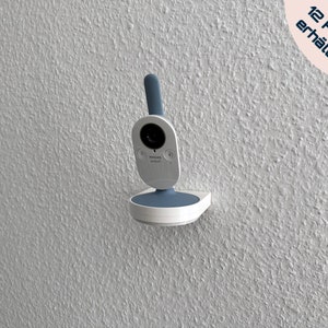Könnte beinhalten: Ein weißer Babymonitor mit einem blauen Objektiv und einem blauen Sockel. Der Monitor ist an einer weißen Wand montiert. Der Text "In 12 Farben erhältlich" ist in der oberen rechten Ecke des Bildes sichtbar.