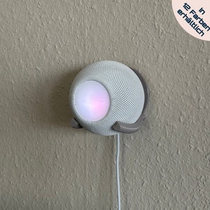 Könnte beinhalten: Ein weißer intelligenter Lautsprecher mit einem hellrosa und violetten Leuchten, der an einer Wand mit einer grauen Halterung befestigt ist. Der Lautsprecher hat ein Mesh-Gitter und einen kleinen Knopf an der Vorderseite. Der Text "In 12 Farben erhältlich" ist in der oberen rechten Ecke des Bildes sichtbar.