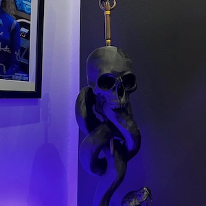 Könnte beinhalten: Eine dunkelgraue Skulptur, die einen Schädel mit einer sich darum windenden Schlange zeigt. Das Maul der Schlange ist geöffnet und zeigt ihre Zähne. Die Skulptur hat einen langen, verzierten Griff mit einem Schädel an der Spitze. Der Hintergrund ist eine dunkle Wand.