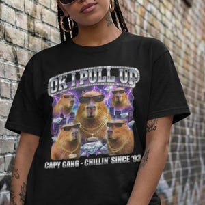 May include: Black t-shirt featuring a graphic of capybaras wearing sunglasses and gold chains, with the text "OK I PULL UP" and "CAPY GANG - CHILLIN' SINCE '93". The shirt is worn by a person in a casual setting.