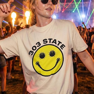Puede incluir: Una persona con una camiseta color crema con el texto "303 STATE" y una carita sonriente amarilla. La persona lleva gafas de sol redondas y está en un concierto al aire libre con luces láser.