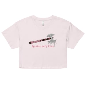 May include: A light pink crop top featuring a graphic of a silver axe with a brown handle wrapped in pink string. The text "Handle with Care" is printed below the axe in a gothic font.