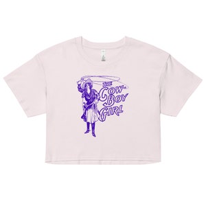 Könnte beinhalten: Ein kurzes T-Shirt in Lila und Weiß mit einer Vintage-Illustration einer Frau, die einen Cowboyhut trägt und ein Lasso hält. Der Text "THE COWBOY GIRL" ist in Lila auf dem Shirt gedruckt.
