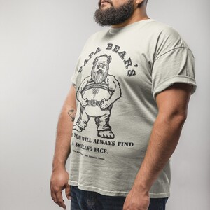 May include: A man wearing a light green t-shirt with a black and white illustration of a bear on the front. The shirt says "Papa Bear's" in a circle around the bear. The text below the bear says "You will always find a smiling face."