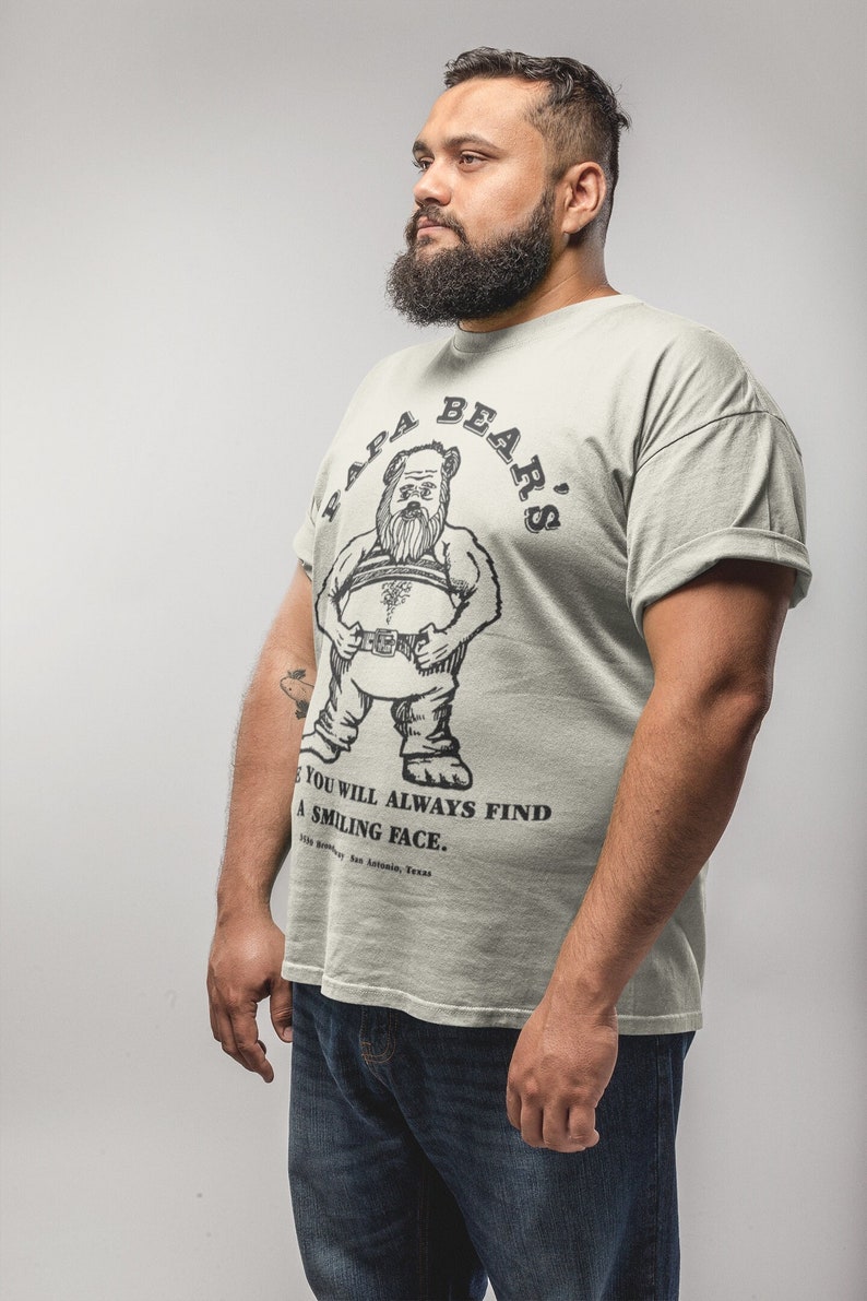 May include: A man wearing a light green t-shirt with a black and white illustration of a bear on the front. The shirt says "Papa Bear's" in a circle around the bear. The text below the bear says "You will always find a smiling face."